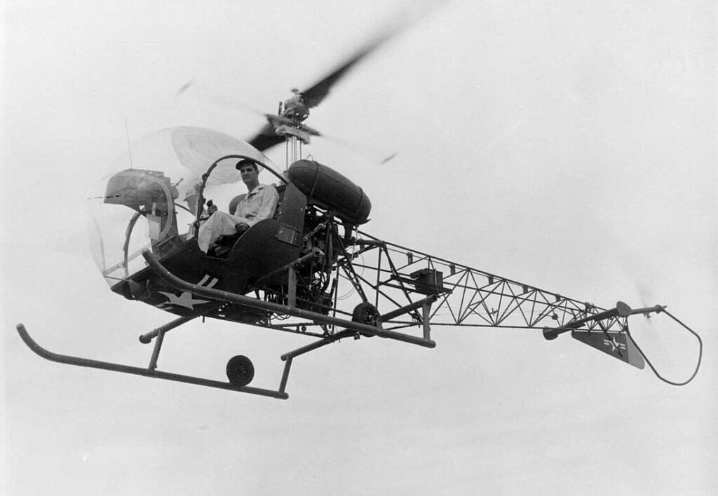  Um helicóptero de estrutura aberta com um único piloto está no ar, visto de lado contra um céu claro. 