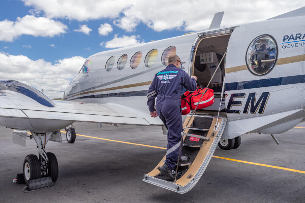 Um homem de uniforme azul, que trabalha com resgate médico em avião, com uma bolsa vermelha na mão, subindo as escadas de um avião branco.