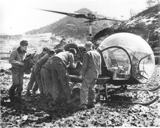 Uma fotografia em preto e branco mostra um grupo de soldados carregando uma maca com uma pessoa ferida para um helicóptero. O helicóptero, com um design de bolha, está estacionado em um terreno lamacento, com montanhas ao fundo.